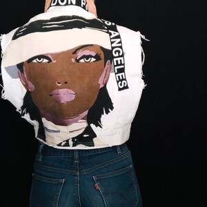May include: A white denim jacket with a black and white graphic of a woman's face and the text "Los Angeles" on the back. The jacket has distressed details and a cropped fit.