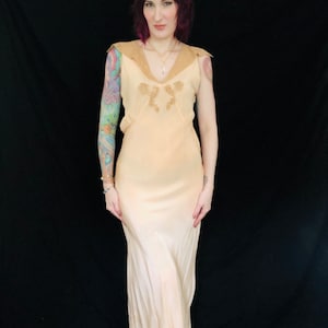 May include: A pale peach colored satin slip dress with a lace trim neckline and a fitted bodice. The dress has a flowing skirt and is sleeveless.
