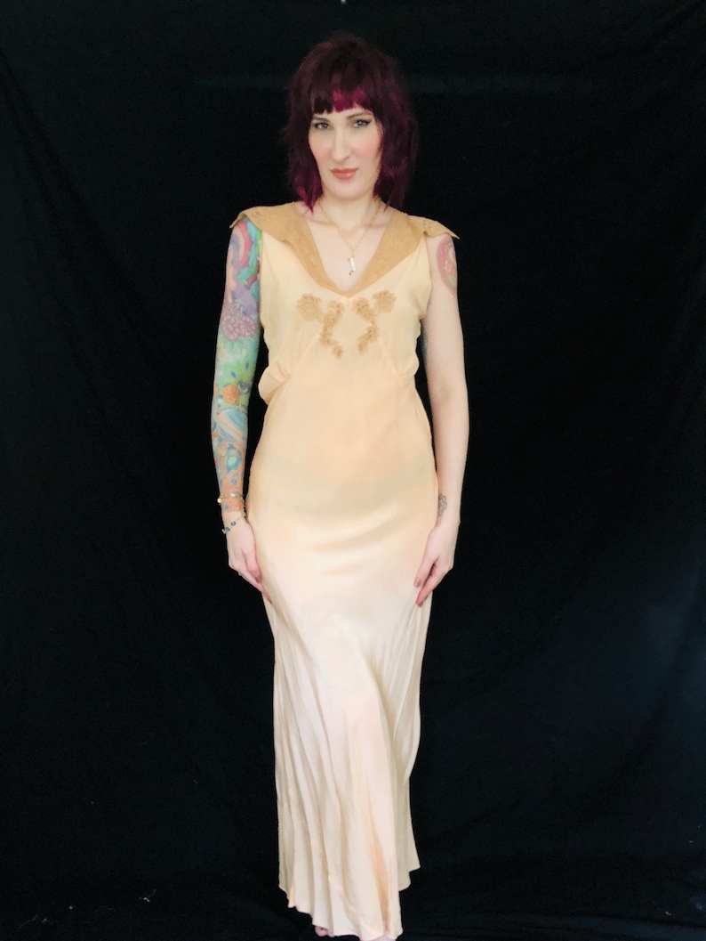 May include: A pale peach colored satin slip dress with a lace trim neckline and a fitted bodice. The dress has a flowing skirt and is sleeveless.