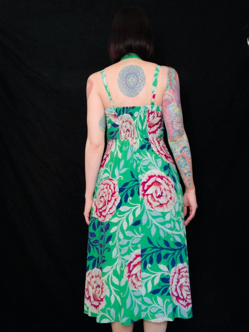 May include: A green floral print dress with a halter neckline and spaghetti straps. The dress features large pink and white peony-like flowers and blue leaves on a green background. The dress is knee-length.