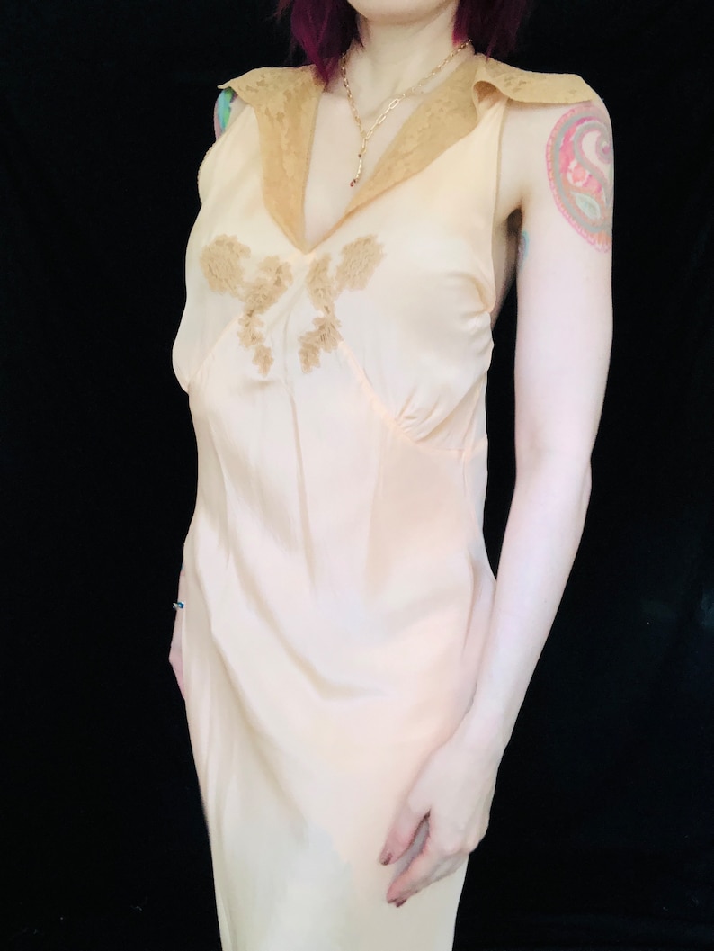 May include: A pale pink silk slip dress with a lace trim collar and delicate floral embroidery. The dress has a fitted bodice and a flowing skirt.