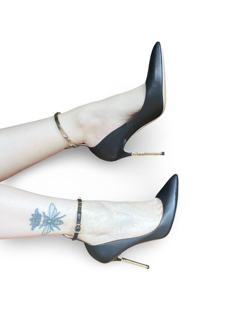 May include: Black high-heeled shoes with pointed toes and ankle straps. The heels have a gold-colored chain detail. The shoes are shown on a white background.