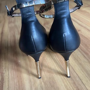 May include: A pair of black leather heeled sandals. The ankle straps are adorned with gold-colored square studs. The heels are thin and gold-colored, with a textured design. The sandals are on a wooden surface.