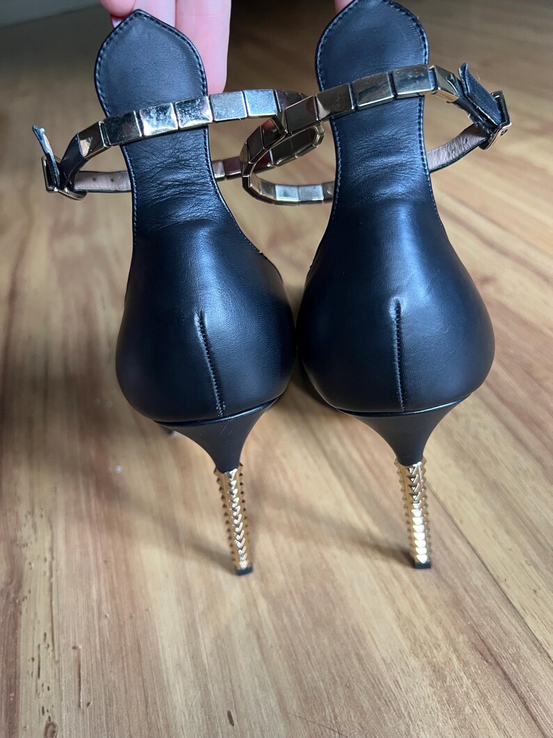May include: A pair of black leather heeled sandals. The ankle straps are adorned with gold-colored square studs. The heels are thin and gold-colored, with a textured design. The sandals are on a wooden surface.