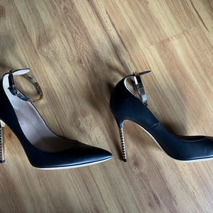 May include: A pair of black high heels with pointed toes. The heels have a gold-colored, studded stiletto heel and a silver ankle strap. The shoes are set against a wooden floor.