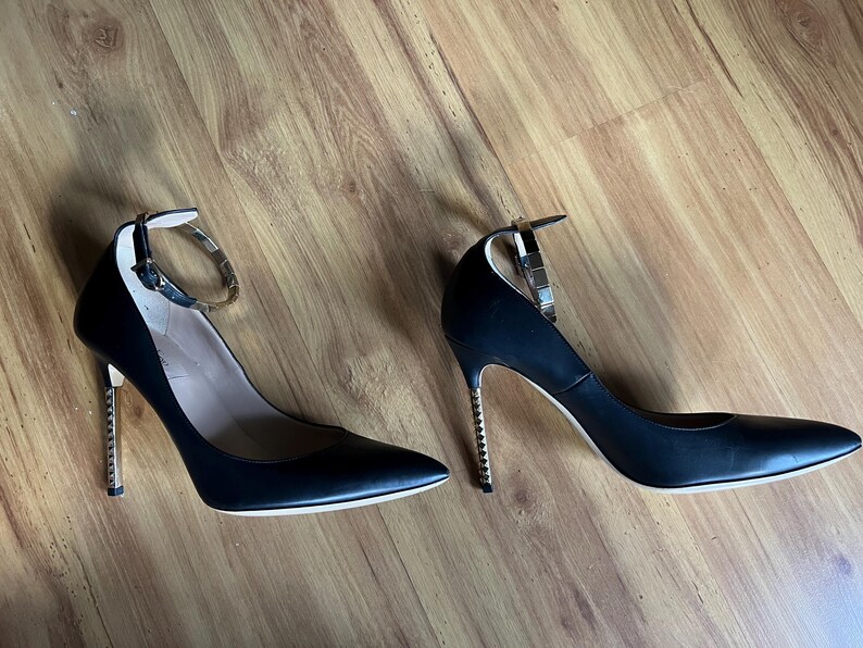 May include: A pair of black high heels with pointed toes. The heels have a gold-colored, studded stiletto heel and a silver ankle strap. The shoes are set against a wooden floor.