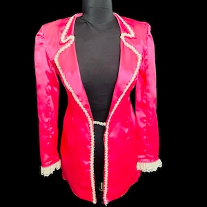 May include: A vibrant pink satin jacket with white lace trim along the lapels, cuffs, and front edges. The jacket is displayed on a black mannequin against a black background. The jacket has a tailored fit and a classic design.