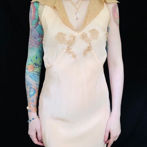 May include: A sleeveless, peach-colored slip dress with a lace trim collar and delicate floral embroidery. The dress has a fitted bodice and a flowing skirt.