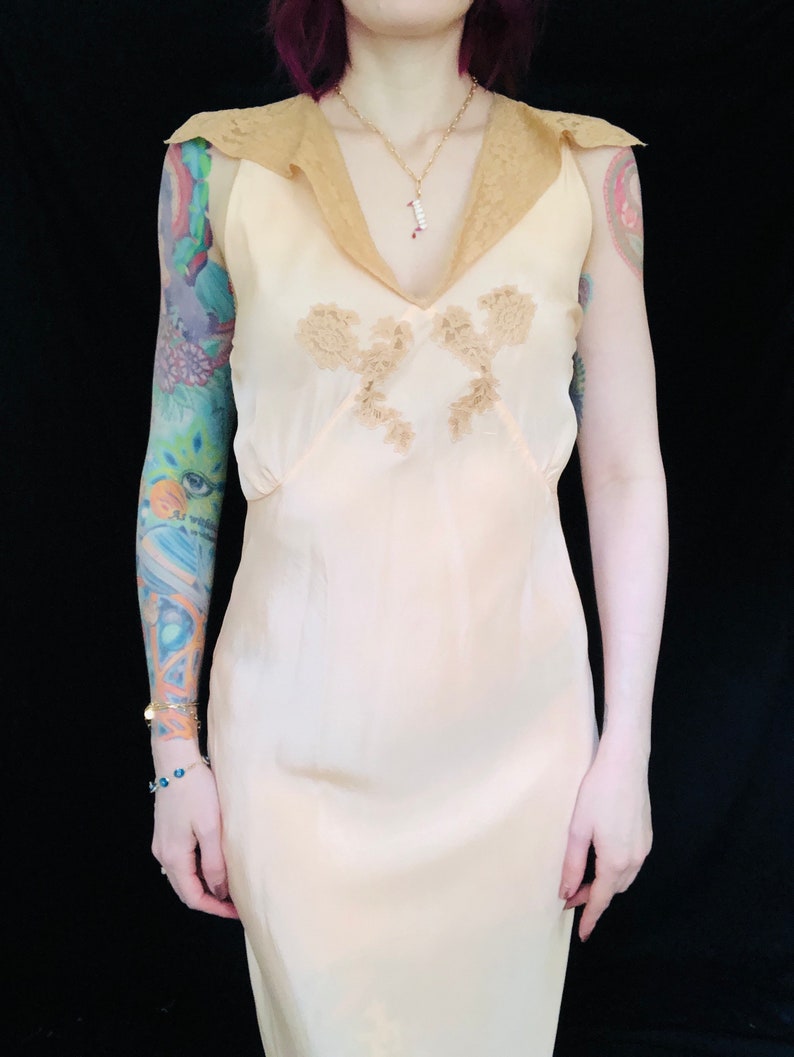 May include: A sleeveless, peach-colored slip dress with a lace trim collar and delicate floral embroidery. The dress has a fitted bodice and a flowing skirt.