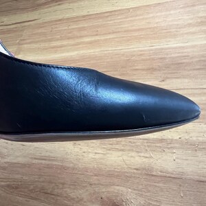 May include: Close-up of a black leather shoe with a pointed toe. The shoe has a smooth, shiny surface and a thin sole. The heel is not visible in the image. The shoe is set against a wooden background.