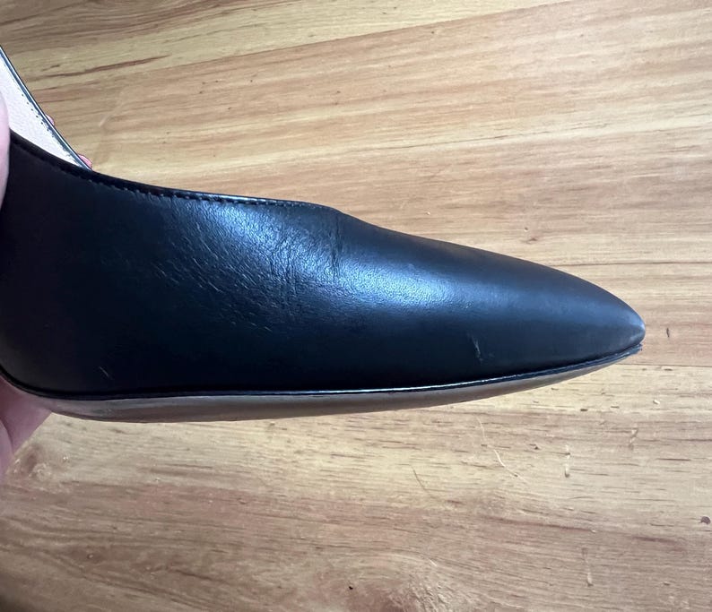 May include: Close-up of a black leather shoe with a pointed toe. The shoe has a smooth, shiny surface and a thin sole. The heel is not visible in the image. The shoe is set against a wooden background.