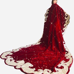 以下が含まれることがあります： ダイヤモンドステッチの模様とクリーム色の装飾的な縁取りが施された、長く流れるような赤いベルベットのケープ。ケープは長いトレーンが特徴で、小さく輝くアクセントで飾られています。