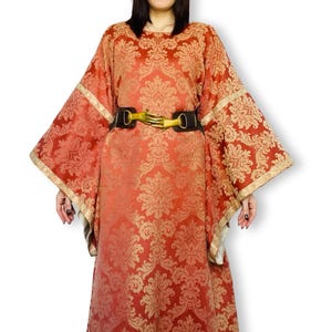 May include: A floor-length orange and gold damask gown with wide, flared sleeves and gold trim. The dress is cinched at the waist with a black belt featuring a gold-colored buckle shaped like two hands meeting in the center, creating a distinct focal point.