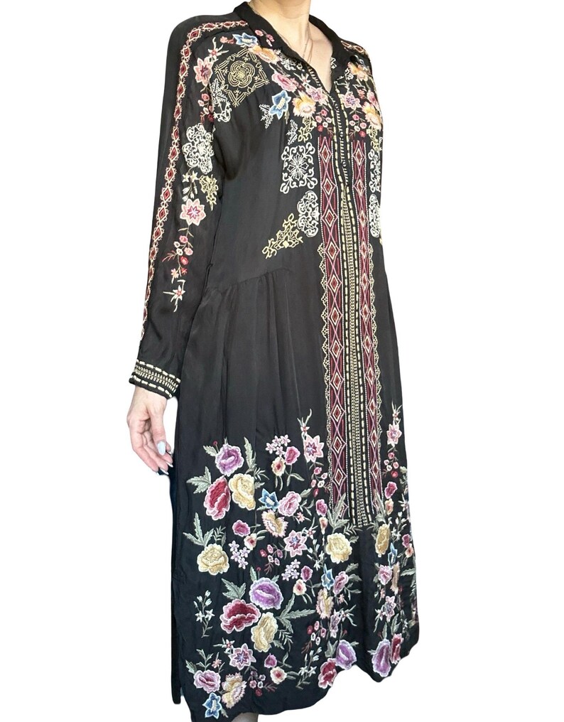 May include: A black long-sleeved dress featuring a vibrant floral embroidery pattern in shades of pink, purple, yellow, and white. The garment includes a central vertical panel with geometric diamond motifs and a collared neckline with a V-shaped opening.