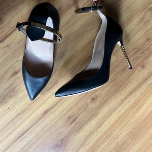 May include: A pair of black high-heeled shoes with pointed toes and ankle straps. The heels have a gold-colored metal design. One shoe has a gold-colored metal strap across the top of the foot.