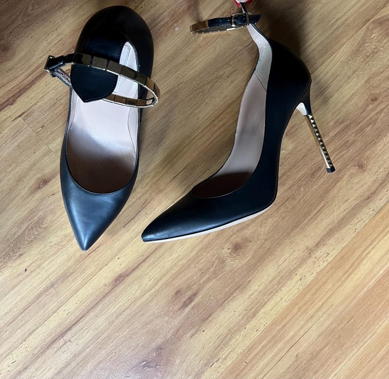 May include: A pair of black high-heeled shoes with pointed toes and ankle straps. The heels have a gold-colored metal design. One shoe has a gold-colored metal strap across the top of the foot.