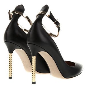 May include: Black leather high heels with gold-tone studded heels and ankle straps. The shoes have a pointed toe and a gold-tone buckle closure. The heels are approximately 4 inches tall.