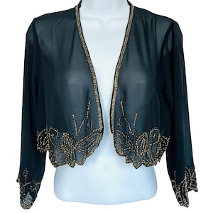 Peut inclure: Boléro noir transparent à manches longues. La veste présente une bordure perlée dorée et des motifs floraux perlés dorés complexes sur le devant et les manches. Le boléro est présenté sur un mannequin blanc.