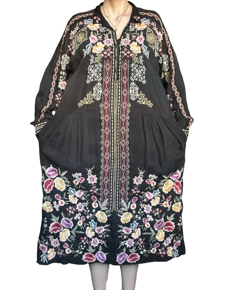 May include: A black long-sleeved dress featuring a central vertical panel with geometric patterns and colorful floral embroidery at the hem and shoulders. The garment has a button-down collar, long sleeves, and a loose, flowing silhouette.