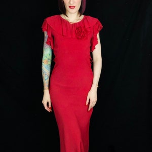 May include: A red midi dress with a ruffled neckline and a flower accent. The dress has short sleeves and a fitted bodice.