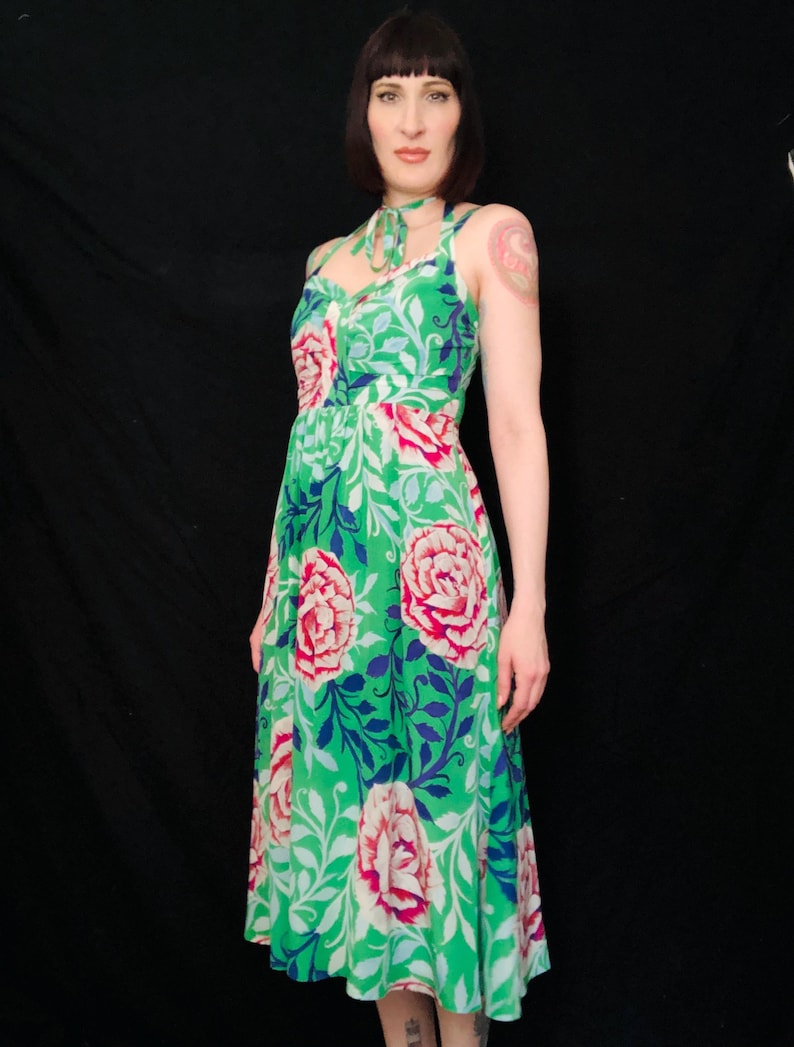 May include: A green floral print dress with a halter neckline and a flowing skirt. The dress features large pink and white flowers with blue and white foliage against a bright green background. The dress is sleeveless and appears to be made of a lightweight fabric.