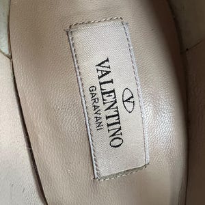 May include: Close-up of the interior of a shoe, showcasing a label with the brand name "VALENTINO GARAVANI" in black text. The label is rectangular with a white background and a silver border. The Valentino logo is also visible.