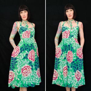 May include: A green halter dress with a floral pattern featuring large pink roses, blue leaves, and white accents. The dress has a fitted bodice, a flared skirt, and pockets. The model is wearing the dress.