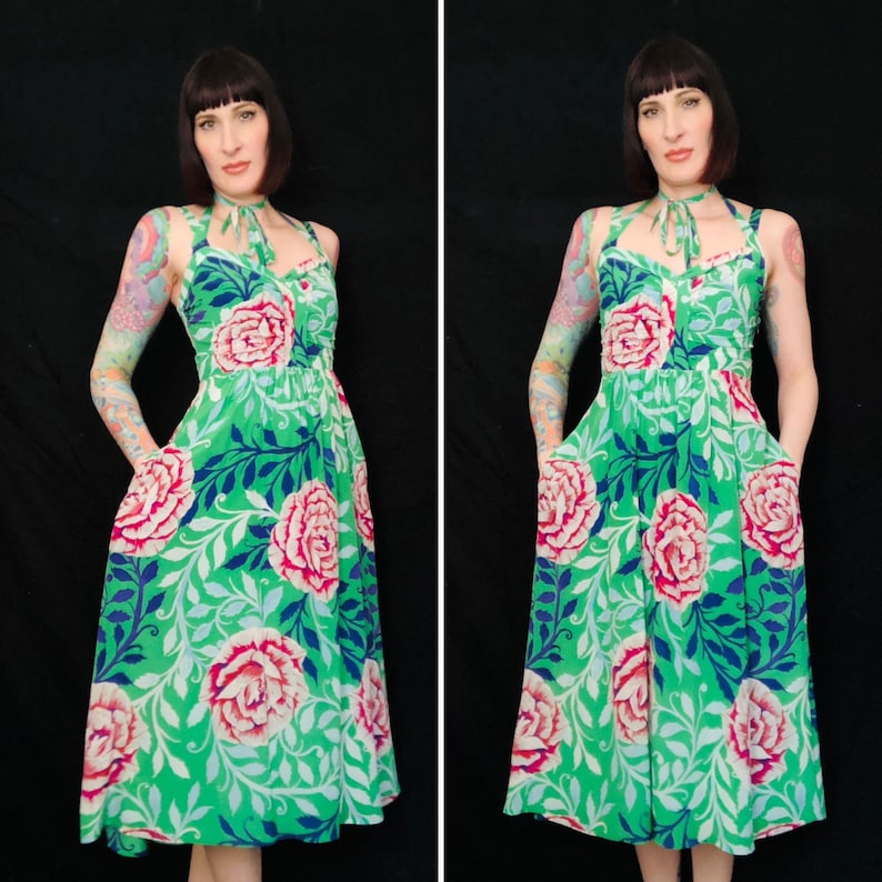 May include: A green halter dress with a floral pattern featuring large pink roses, blue leaves, and white accents. The dress has a fitted bodice, a flared skirt, and pockets. The model is wearing the dress.