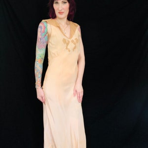 May include: A woman wearing a peach-colored slip dress with delicate lace detailing. The dress has a V-neckline and sleeveless design. The woman has pink hair and is standing against a black background.