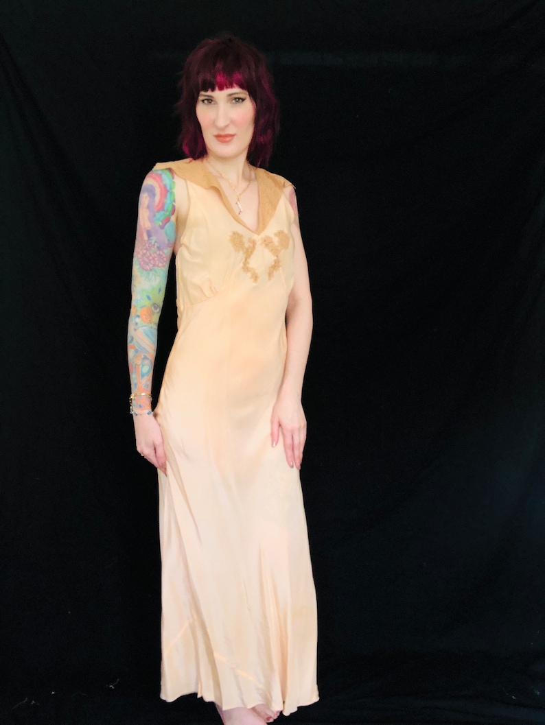 May include: A woman wearing a peach-colored slip dress with delicate lace detailing. The dress has a V-neckline and sleeveless design. The woman has pink hair and is standing against a black background.
