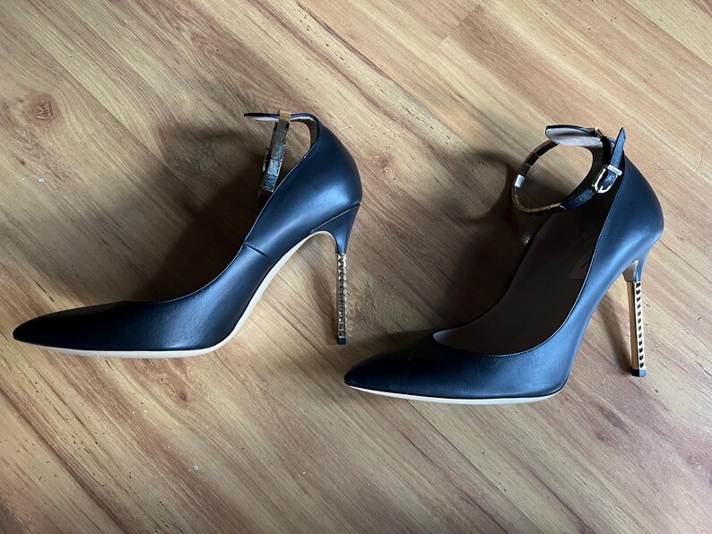 May include: A pair of black leather high heels with pointed toes and ankle straps. The heels have a gold-colored, textured design. The shoes are set against a wooden background.