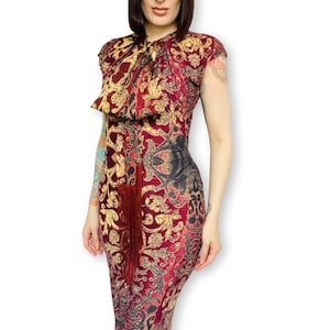 Puede incluir: Vestido hasta la rodilla con un estampado floral burdeos, dorado y negro. El vestido presenta un cuello alto con un lazo y mangas cortas. Una larga borla burdeos cuelga de la cintura. El vestido es entallado.