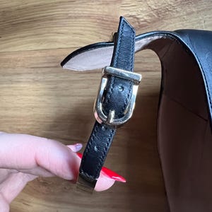 May include: Close-up of a black leather shoe with a gold-colored buckle and strap. The shoe has a high heel and a strap that fastens around the ankle. The buckle is rectangular and has a small pin.