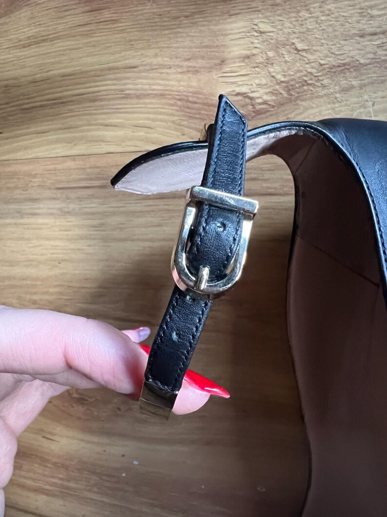 May include: Close-up of a black leather shoe with a gold-colored buckle and strap. The shoe has a high heel and a strap that fastens around the ankle. The buckle is rectangular and has a small pin.