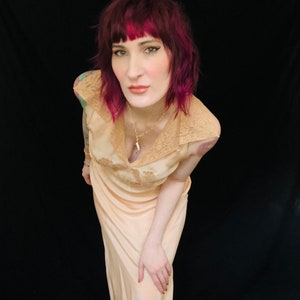 May include: A woman with pink hair wearing a peach-colored satin slip dress with lace trim. The dress has a V-neckline and a fitted bodice. She is wearing a silver necklace with a pendant.