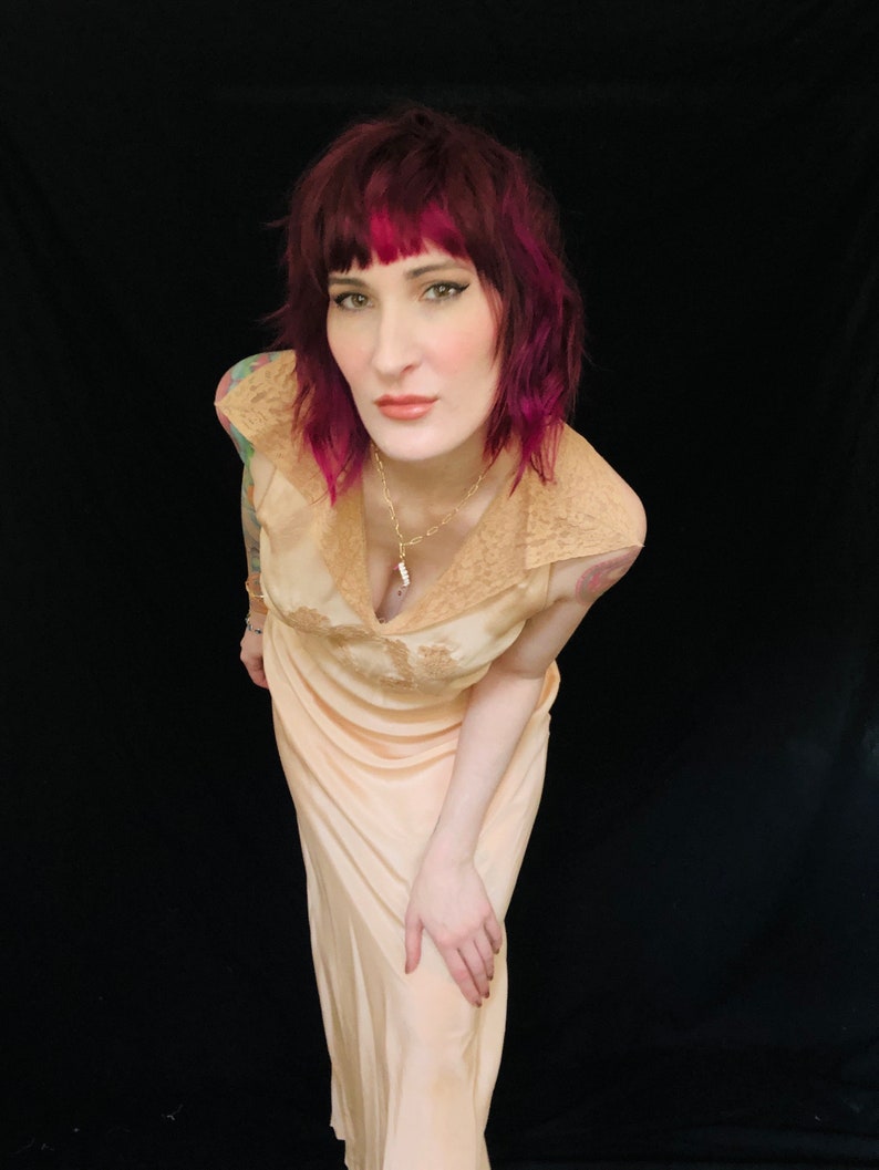 May include: A woman with pink hair wearing a peach-colored satin slip dress with lace trim. The dress has a V-neckline and a fitted bodice. She is wearing a silver necklace with a pendant.