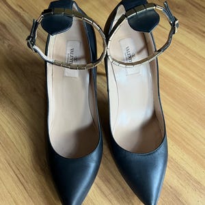 May include: A pair of black leather high heels with pointed toes. The shoes have ankle straps with gold-colored metal accents and a gold-colored metal band around the ankle. The brand name "VALENTINO GARAVANI" is printed on the insole.