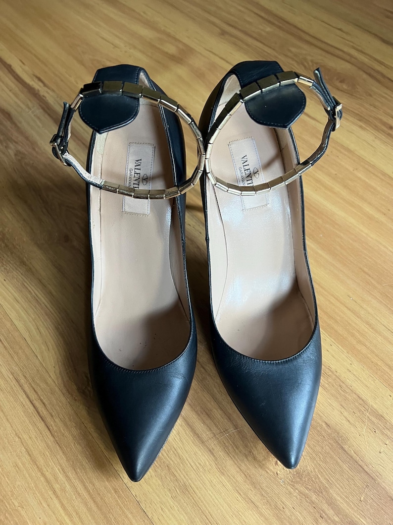 May include: A pair of black leather high heels with pointed toes. The shoes have ankle straps with gold-colored metal accents and a gold-colored metal band around the ankle. The brand name "VALENTINO GARAVANI" is printed on the insole.