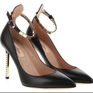 May include: A pair of black leather high heels with pointed toes. The heels have gold-colored metal accents on the heel and ankle strap. The ankle strap is adorned with gold-colored square studs. The brand name "Valentino" is visible on the insole.