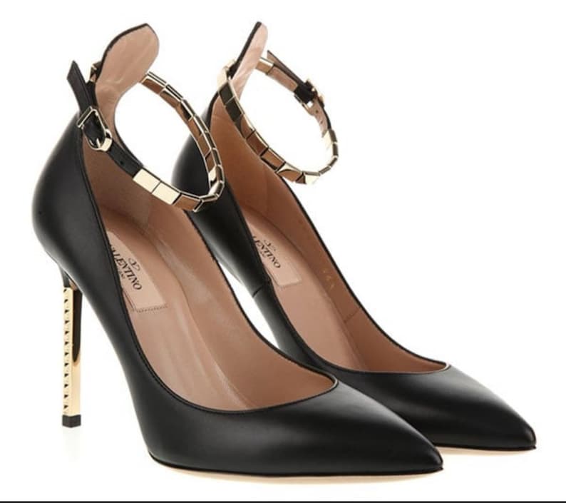 May include: A pair of black leather high heels with pointed toes. The heels have gold-colored metal accents on the heel and ankle strap. The ankle strap is adorned with gold-colored square studs. The brand name "Valentino" is visible on the insole.