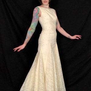 May include: A woman wearing a white lace gown with a fitted bodice and a flowing skirt. The gown is adorned with small, sparkling embellishments. The woman has dark hair and is standing against a black background.