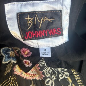 May include: A black garment featuring a white rectangular label with the text BIYA JOHNNY WAS in black and red, and a smaller white tag indicating size M and Made in China. The fabric is decorated with intricate floral embroidery in pink, blue, and gold.