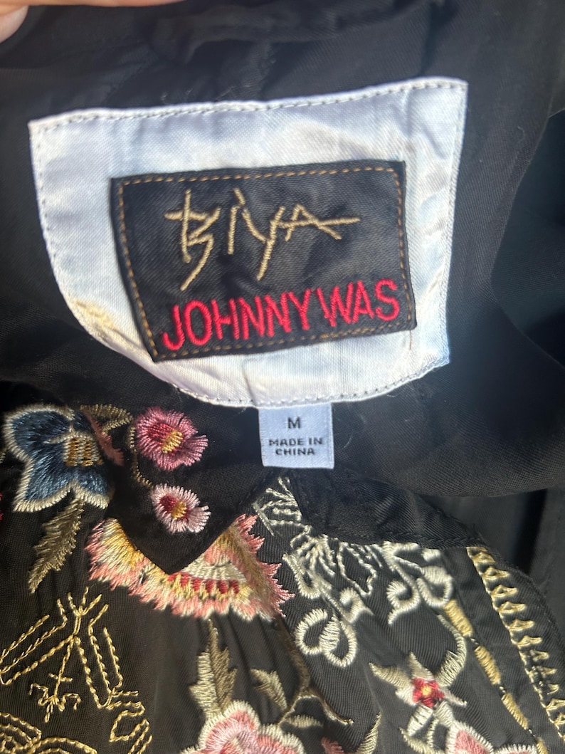 May include: A black garment featuring a white rectangular label with the text BIYA JOHNNY WAS in black and red, and a smaller white tag indicating size M and Made in China. The fabric is decorated with intricate floral embroidery in pink, blue, and gold.