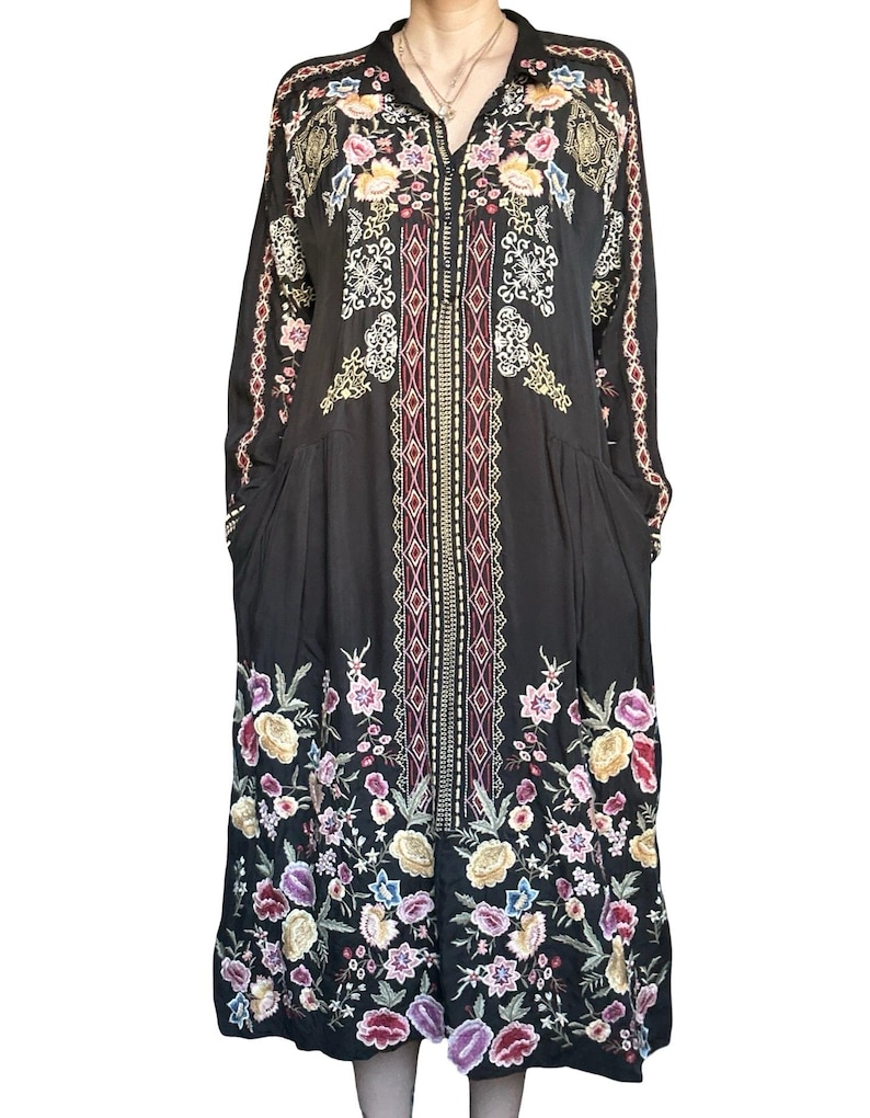 May include: A black long-sleeved midi dress featuring a central vertical panel with geometric patterns and colorful floral embroidery. The garment includes a button-down front, a collar, and intricate flower motifs in shades of pink, yellow, and blue.