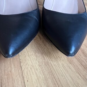 May include: Close-up of two black leather pointed-toe pumps. The shoes have a glossy finish and are set against a wooden floor. The heels are not visible in the image.