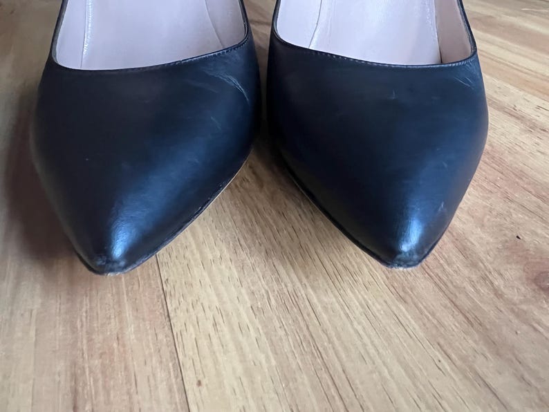 May include: Close-up of two black leather pointed-toe pumps. The shoes have a glossy finish and are set against a wooden floor. The heels are not visible in the image.