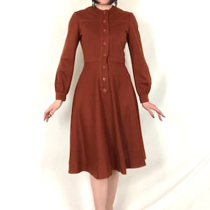 May include: A solid rust-coloured midi dress featuring long sleeves with buttoned cuffs, a high round neckline, and a central vertical button-down placket extending from the collar to the waistline, finished with a flared skirt that falls below the knees.