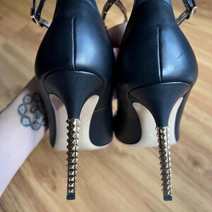 May include: Pair of black leather high heels with gold-colored studded heels and ankle straps. The heels have a stiletto design and are approximately 4 inches (10 cm) tall. The ankle straps have a gold-colored chain detail.