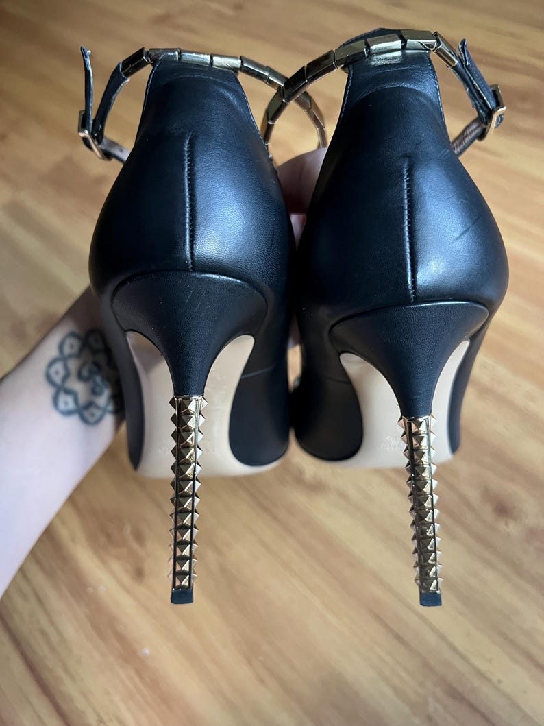 May include: Pair of black leather high heels with gold-colored studded heels and ankle straps. The heels have a stiletto design and are approximately 4 inches (10 cm) tall. The ankle straps have a gold-colored chain detail.