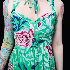 May include: A green halter dress with a floral pattern featuring large pink and white flowers, blue accents, and white leaves. The dress has a tie at the neck and a fitted bodice. The fabric is a vibrant green.